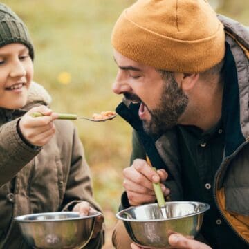 A child in a green knit hat and brown jacket feeds a spoonful of food to a bearded adult wearing an orange beanie and gray jacket. Both hold metal bowls and are sitting outdoors. The background is blurred, showing greenery.