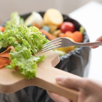A person uses a fork to scrape fresh vegetable scraps, including lettuce, cucumbers, and carrot peels, from a wooden cutting board into a garbage bin lined with a black trash bag.