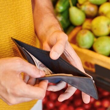 A person wearing a yellow shirt holds an open wallet containing cash. In the background, there are stacks of green pears and red apples in wooden crates at a market or grocery store.