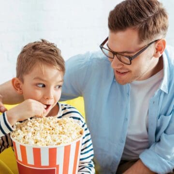 A man with glasses and a boy are sitting on a yellow couch. The boy is eating popcorn from a large striped bucket. The man is smiling at the boy, with his arm resting on the back of the couch. They are indoors.