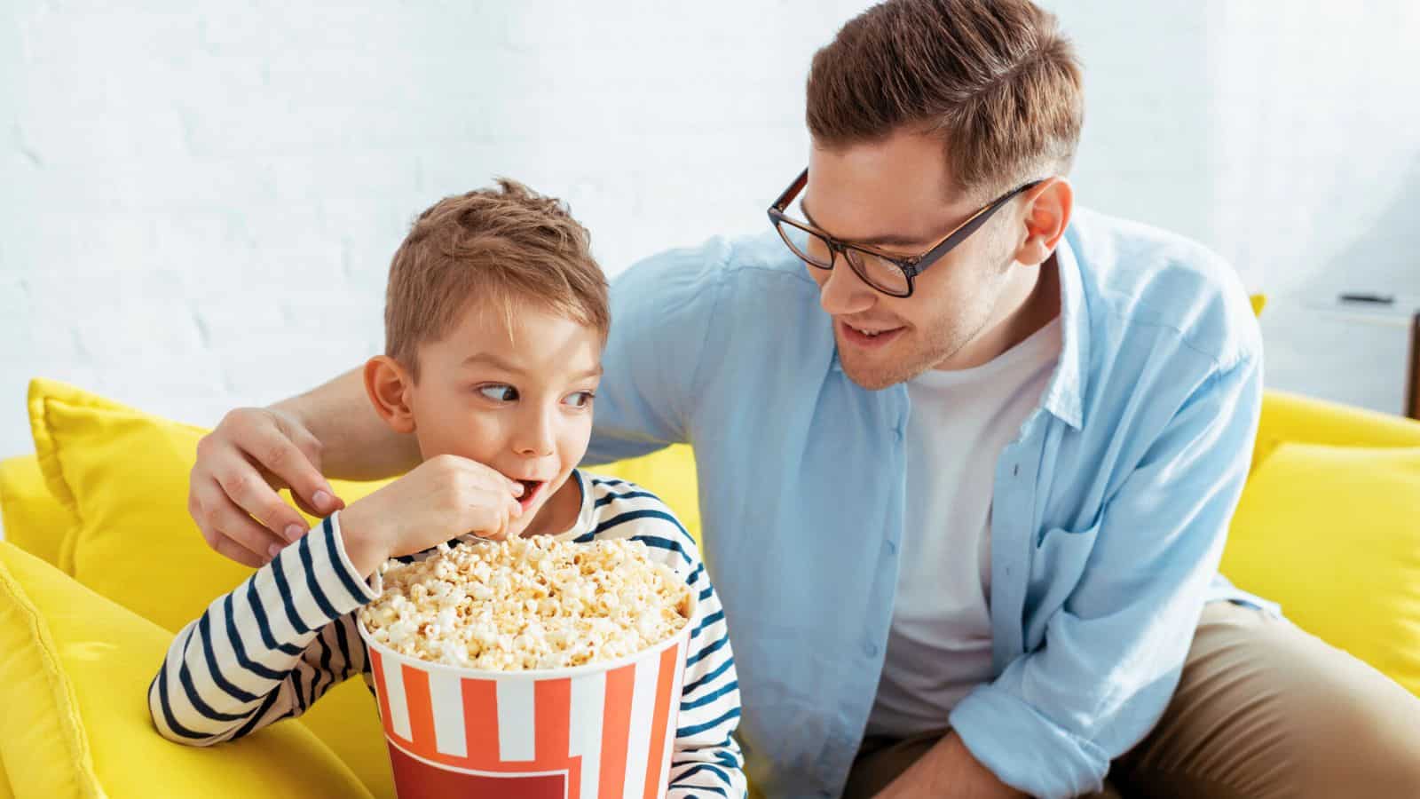 A man with glasses and a boy are sitting on a yellow couch. The boy is eating popcorn from a large striped bucket. The man is smiling at the boy, with his arm resting on the back of the couch. They are indoors.