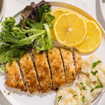 A plate with sliced breaded chicken, garnished with lemon slices and mixed greens. Accompanied by mashed potatoes. A fork is placed nearby on the table, with herbs partially visible.