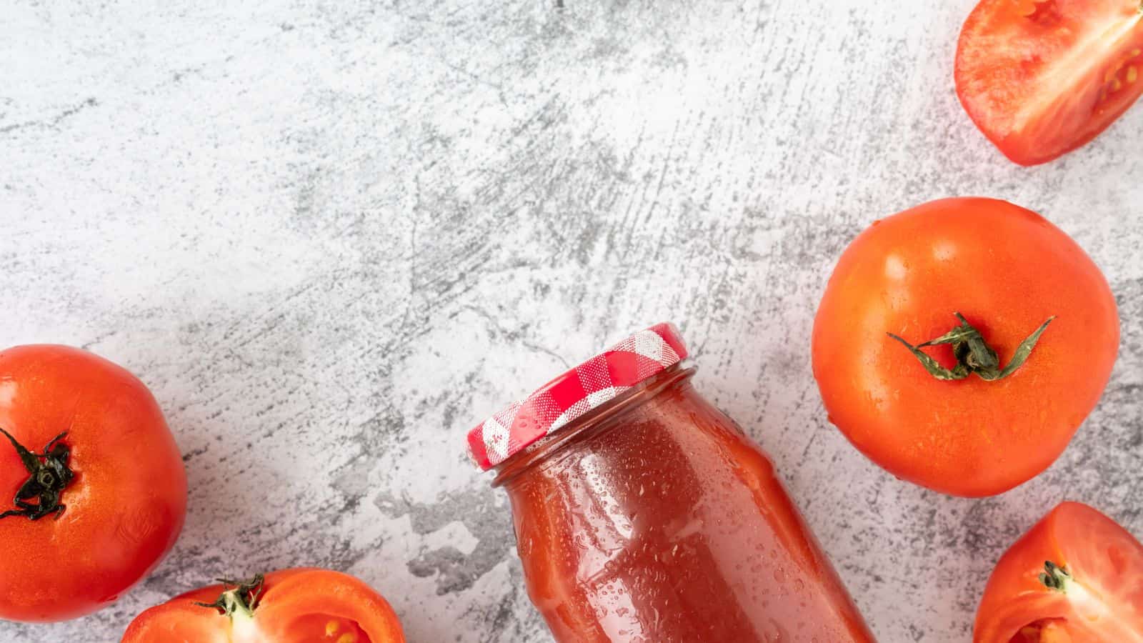 A jar of tomato sauce with a red and white checkered lid is placed on a gray textured surface. Whole and halved tomatoes are arranged around the jar. The background is a light gray, adding contrast to the red tomatoes and jar.