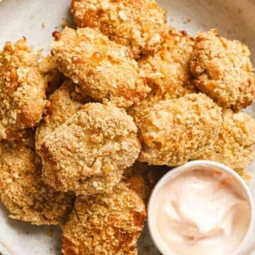 A white bowl holds several pieces of breaded and fried cauliflower, arranged neatly. A small cup of pink dipping sauce sits beside them, perfect for a kid-friendly snack. The background features a light neutral color, creating an inviting setting for parents exploring new recipes.