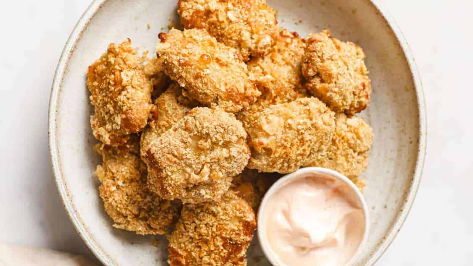 A white bowl holds several pieces of breaded and fried cauliflower, arranged neatly. A small cup of pink dipping sauce sits beside them, perfect for a kid-friendly snack. The background features a light neutral color, creating an inviting setting for parents exploring new recipes.