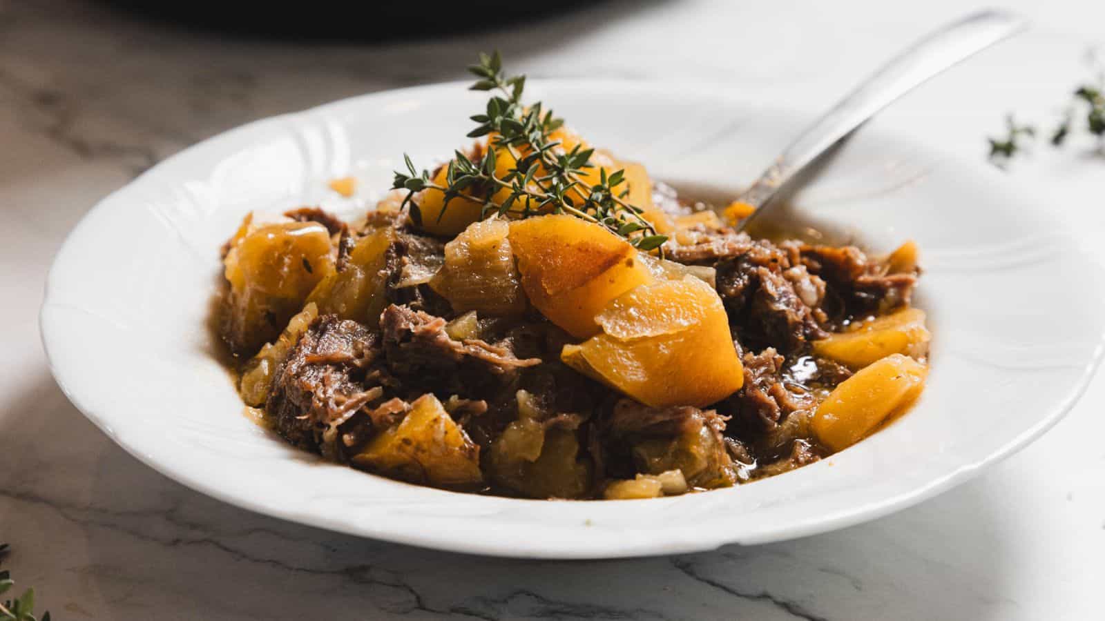 A white plate containing a stew made of tender shredded meat and chunks of potatoes, garnished with sprigs of fresh thyme. A spoon rests on the side of the dish, placed on a marble surface.