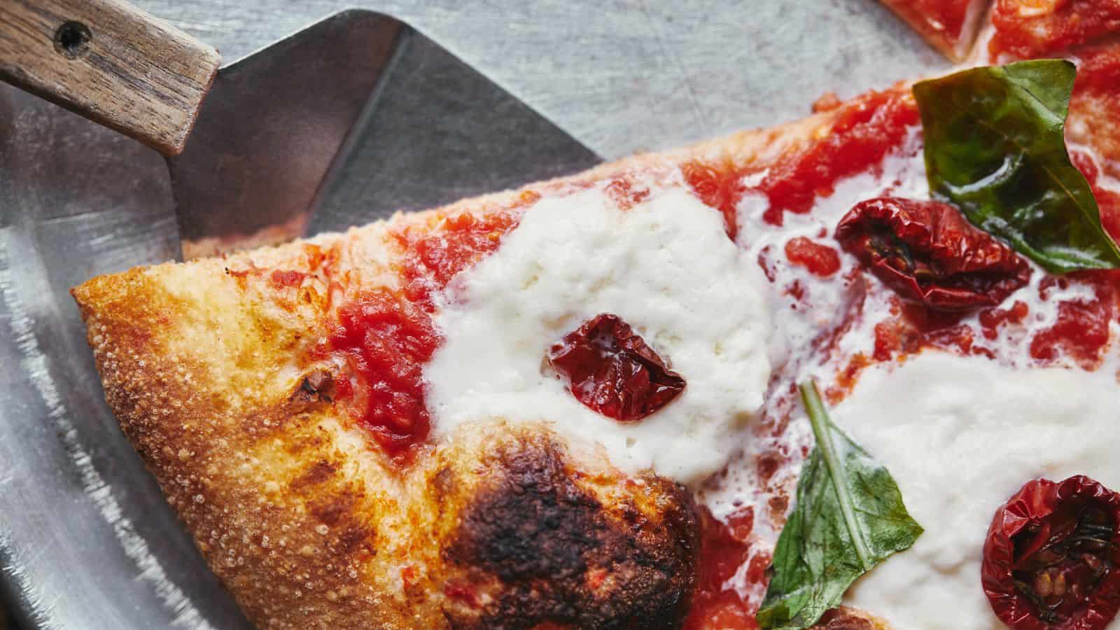 Close-up of a slice of pizza on a metal tray with a wooden-handled spatula partially visible. The pizza has a thick crust, melted white cheese, tomato sauce, sundried tomatoes, and fresh basil leaves.