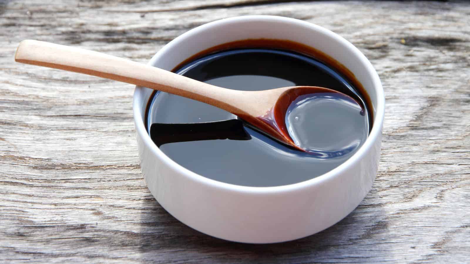 A white ceramic bowl filled with dark soy sauce is placed on a wooden surface. A wooden spoon rests on the edge of the bowl, partially submerged in the soy sauce. The background shows textured wood grain.