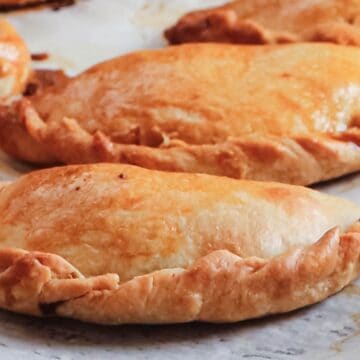 Close-up of golden-brown pastries with crimped edges on a baking sheet. Made from traditional Argentinian empanada dough, they boast a shiny, slightly flaky surface and are arranged in a line. The surface of the baking sheet is lightly browned with spots.