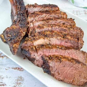 Sliced grilled steak with an unforgettable seasoned crust and visible grill marks on a white rectangular plate. The steak, perfect for a dinner of main courses, features one large bone and is set on a white-washed wooden surface.