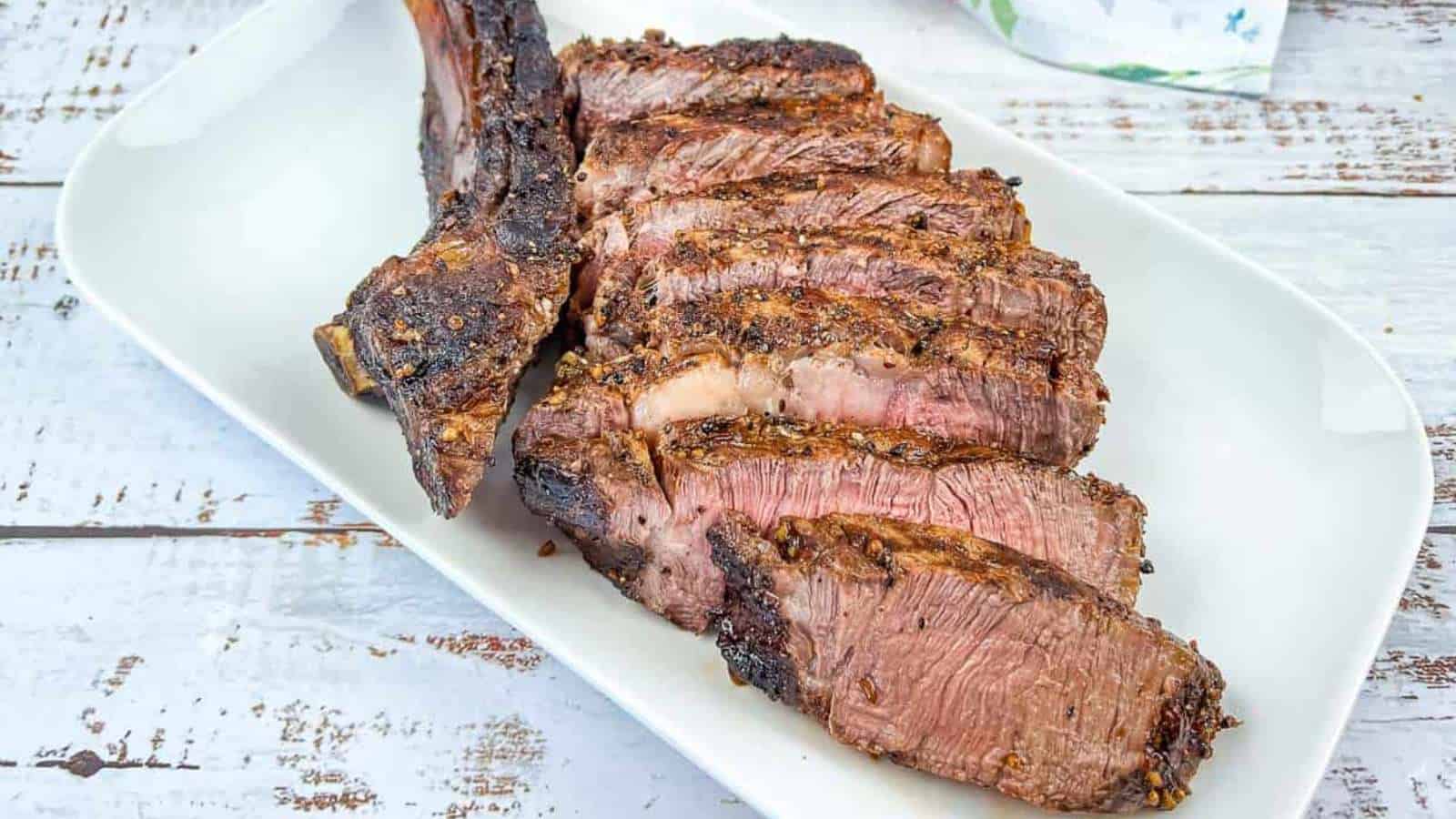 Sliced grilled steak with an unforgettable seasoned crust and visible grill marks on a white rectangular plate. The steak, perfect for a dinner of main courses, features one large bone and is set on a white-washed wooden surface.