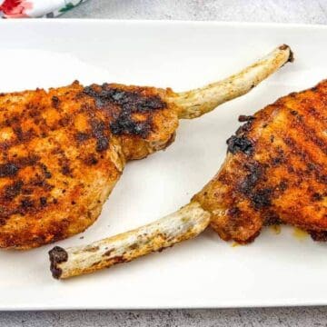 Two grilled pork chops with visible char marks are placed on a white rectangular plate. The chops have bone handles and are seasoned with spices, embodying the spirit of a Backyard BBQ. Lavender flowers and a floral napkin are partially visible in the background.