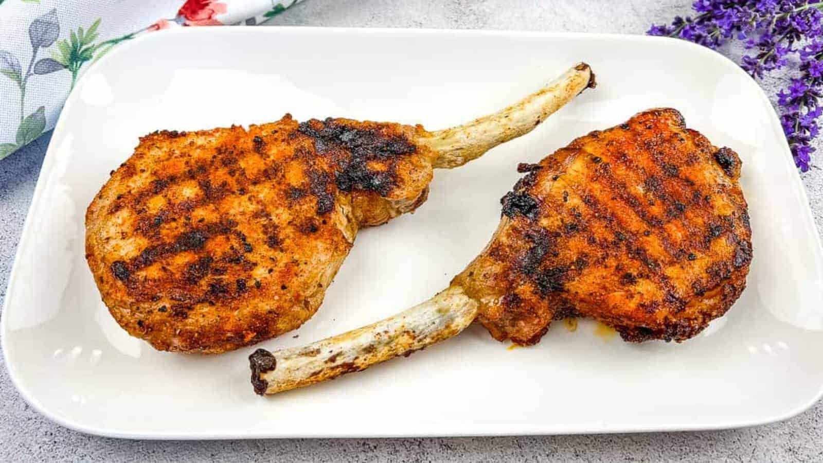 Two grilled pork chops with visible char marks are placed on a white rectangular plate. The chops have bone handles and are seasoned with spices, embodying the spirit of a Backyard BBQ. Lavender flowers and a floral napkin are partially visible in the background.