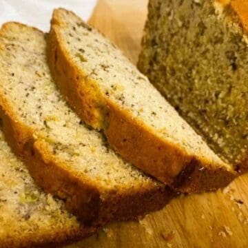 A loaf of homemade banana bread rests on a wooden surface, partially sliced with three pieces leaning against it. The golden-brown crust boasts visible flecks from bananas or nuts. In the background, a white cloth adds a touch of elegance.