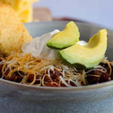 A bowl of chili topped with shredded cheese, a dollop of sour cream, and avocado slices exemplifies easy cooking. A piece of cornbread rests on the bowl's edge, while a blurred background reveals more cornbread and a napkin, perfect for fans of flavorful dishes.