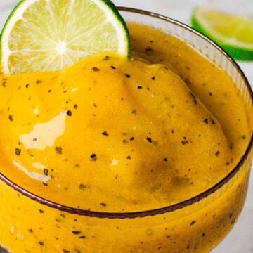 A close-up of a tropical smoothie in a glass, featuring a thick, yellow-orange blend with visible black specks. A slice of lime is placed on the rim. A cut passion fruit and a lime wedge are in the blurred background.