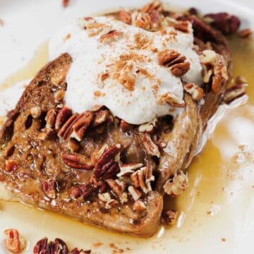 A white plate holds a slice of French toast topped with whipped cream, pecans, and a sprinkle of cinnamon. Syrup is drizzled over the toast, pooling slightly on the plate. The setting is on a light wooden table.