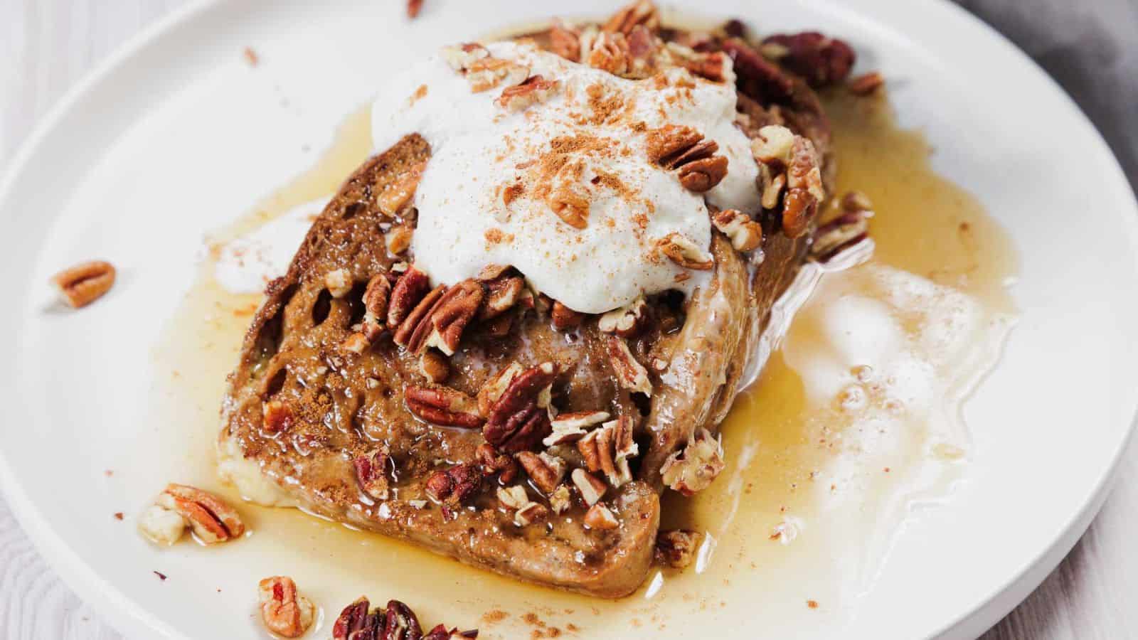 A slice of French toast on a white plate is topped with whipped cream, pecans, a dusting of cinnamon, and drizzled with syrup.