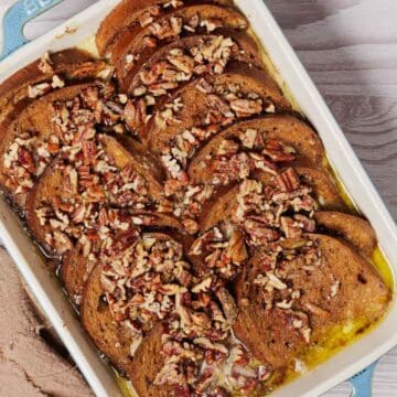 A rectangular baking dish contains sliced French toast, topped with chopped pecans and a drizzle of syrup. A beige cloth is to the left, and a small bowl of cream is partially visible in the upper right corner on a light wooden table.