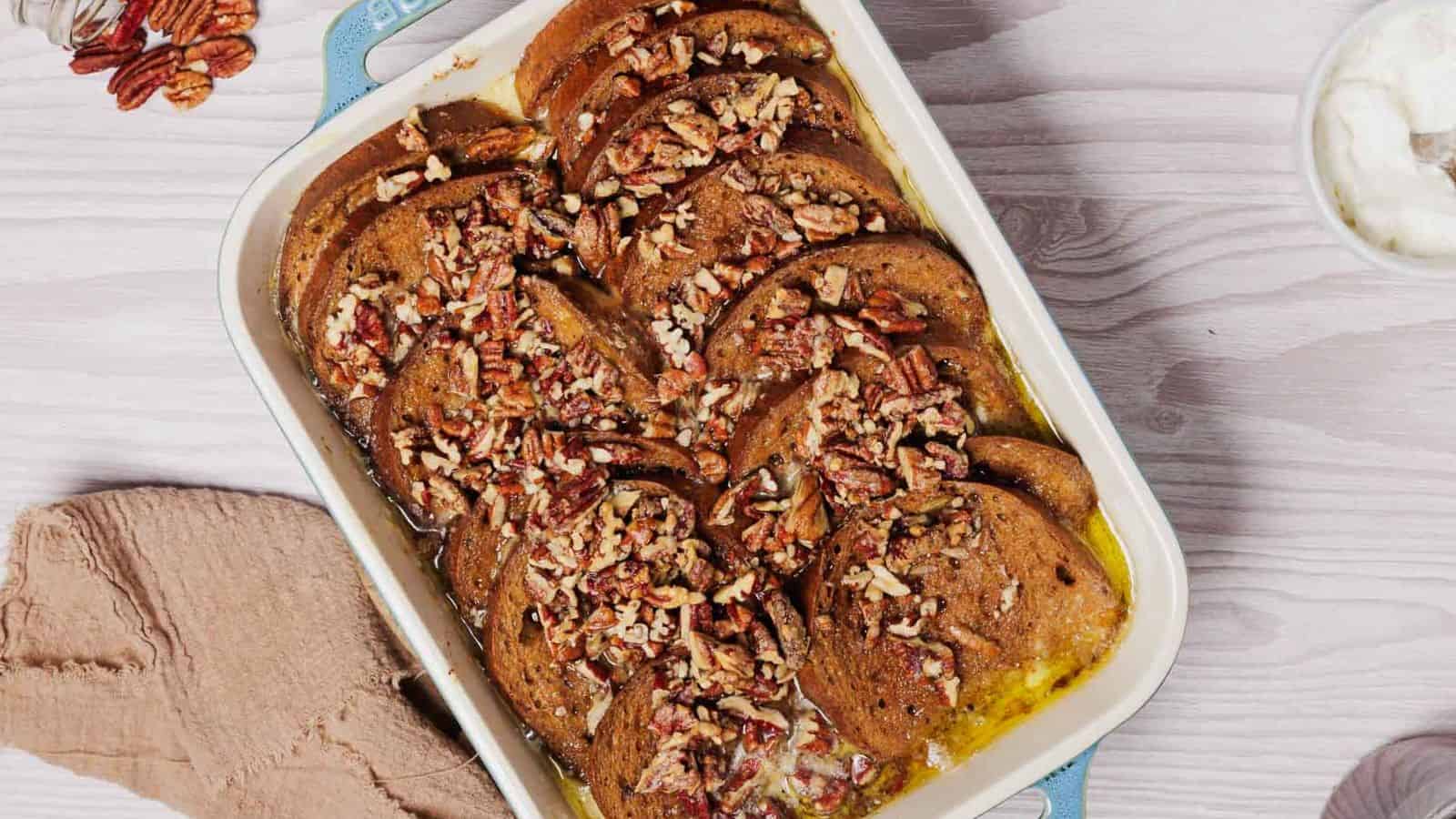 A rectangular baking dish contains sliced French toast, topped with chopped pecans and a drizzle of syrup. A beige cloth is to the left, and a small bowl of cream is partially visible in the upper right corner on a light wooden table.