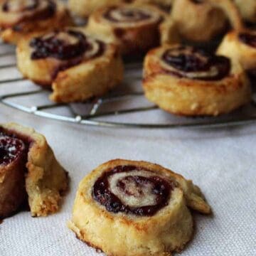 Small pastry rolls filled with fruit jam are arranged on a wire rack and tablecloth. The rolls have a golden-brown crust and swirled appearance, showcasing a rich, dark filling. Some pastries are slightly crumbled around the edges.