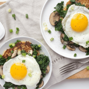 Two plates of breakfast beckon you with fried eggs boasting bright yellow yolks atop sautéed mushrooms and greens. Garnished with chopped green onions, these dishes are perfect breakfast ideas to make you feel like jumping out of bed. A fork rests on the wooden table beside one plate.