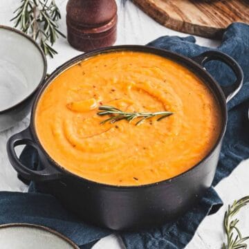 A black pot filled with creamy roasted butternut squash soup, garnished with a sprig of rosemary, sits on a cloth napkin. Nearby lie empty bowls, surrounded by rosemary sprigs and the edge of a wooden board peeking through in the background.