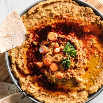 A bowl of roasted carrot hummus garnished with tender carrot slices and a sprig of parsley. The hummus features a swirled design with reddish spices and olive oil on top. Two pieces of flatbread rest beside the bowl on a rustic wooden surface.