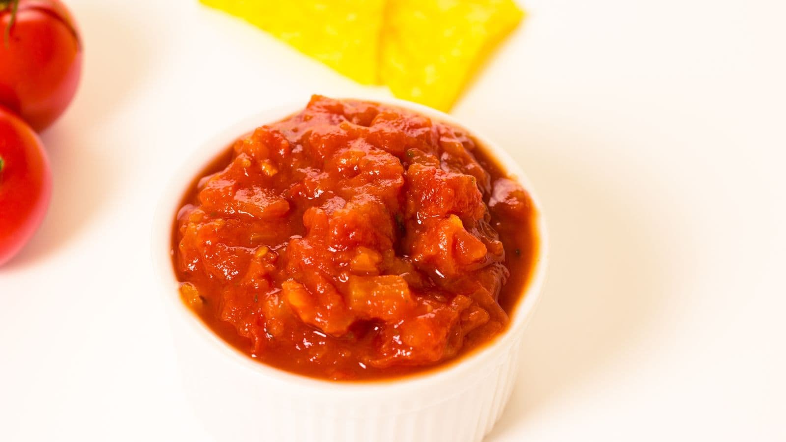 A white ramekin filled with chunky tomato salsa is placed on a white surface. In the background, there are two red tomatoes to the left and two pieces of fresh pineapple to the right.