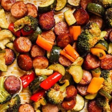 A sheet pan brimming with roasted sliced sausage, broccoli, zucchini, and vibrant bell peppers tempts you to lick the pan clean. A silver spoon peeks from beneath the savory vegetables and meat, all perfectly browned.