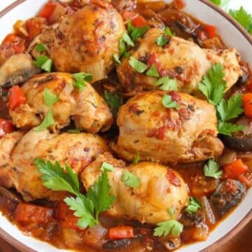 A plate of cooked chicken pieces garnished with parsley. The dish is accompanied by a stew of vegetables including bell peppers and mushrooms. The plate rests on a wooden surface beside a folded napkin.