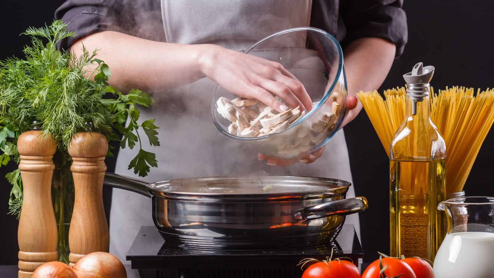 A person wearing an apron adds sliced mushrooms from a glass bowl into a steaming pan on a stove. Nearby are uncooked spaghetti, olive oil, tomatoes, fresh herbs, and wooden pepper grinders.