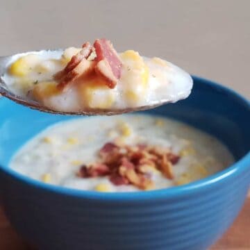 A spoon holding a portion of creamy soup with corn and crumbled bacon is being lifted over a blue bowl filled with the same soup. The bowl is placed on a wooden surface.