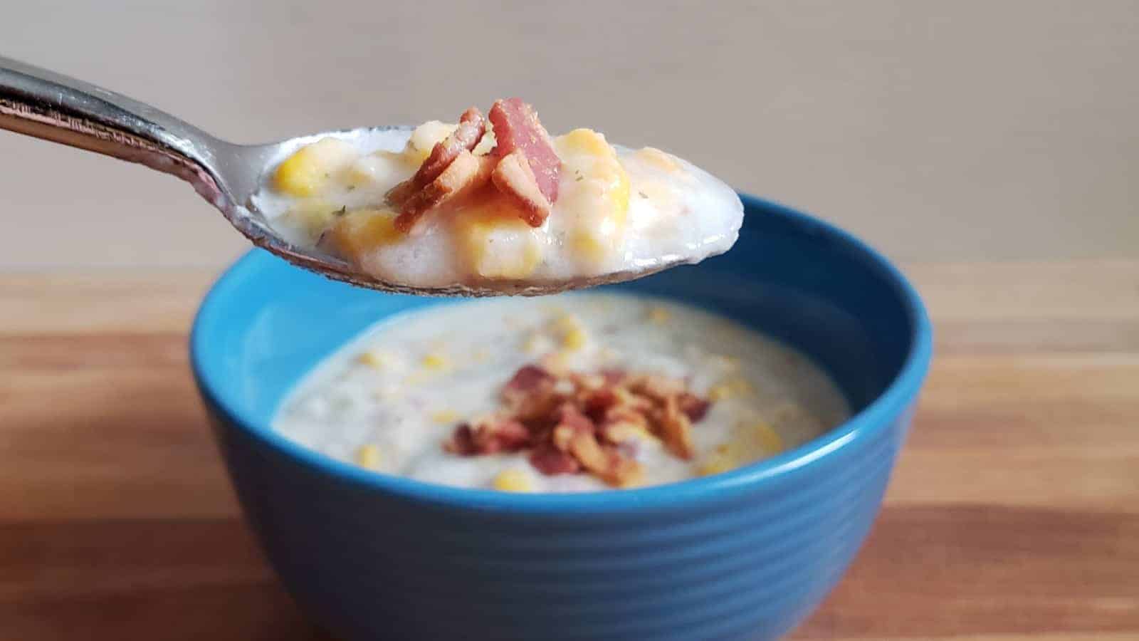 A spoon holding a portion of creamy soup with corn and crumbled bacon is being lifted over a blue bowl filled with the same soup. The bowl is placed on a wooden surface.