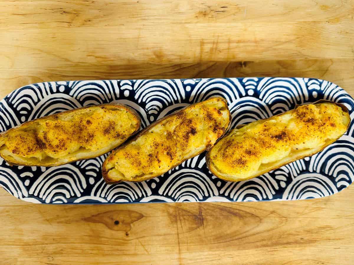 Three baked potato halves topped with melted cheese and seasoning are arranged on a black and white patterned tray, set on a wooden surface.