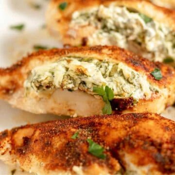 Close-up of stuffed chicken breasts, sliced to show a creamy spinach and cheese filling. The chicken is seasoned with spices and garnished with small parsley pieces, served on a white plate.