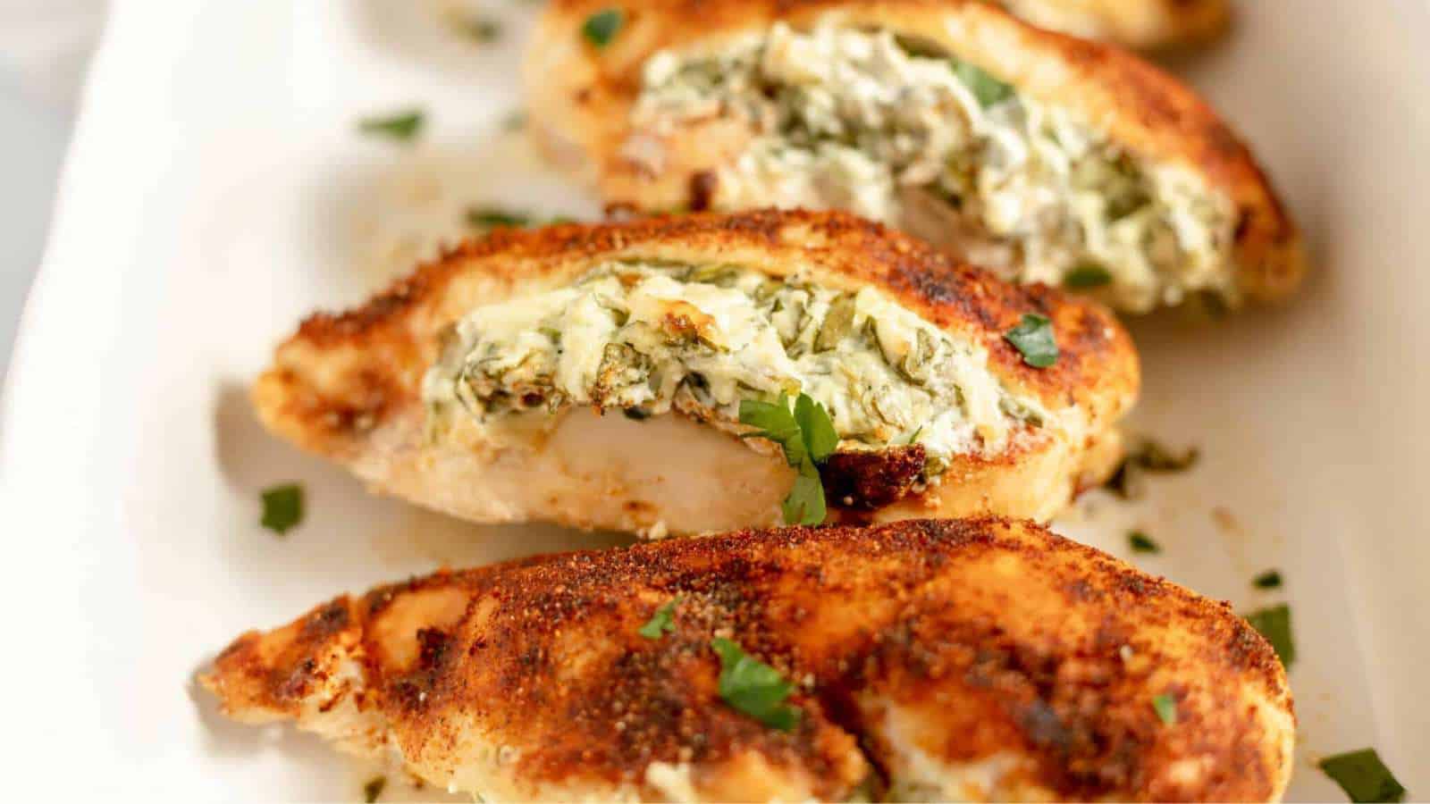 Close-up of stuffed chicken breasts, sliced to show a creamy spinach and cheese filling. The chicken is seasoned with spices and garnished with small parsley pieces, served on a white plate.