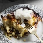 Close-up of a serving of bread pudding on a decorative plate, reminiscent of sweet noodle kugel. The pudding is topped with whipped cream and appears slightly browned on top. A fork is partially inserted into the dish, revealing its soft texture.