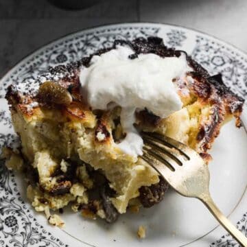 Close-up of a serving of bread pudding on a decorative plate, reminiscent of sweet noodle kugel. The pudding is topped with whipped cream and appears slightly browned on top. A fork is partially inserted into the dish, revealing its soft texture.