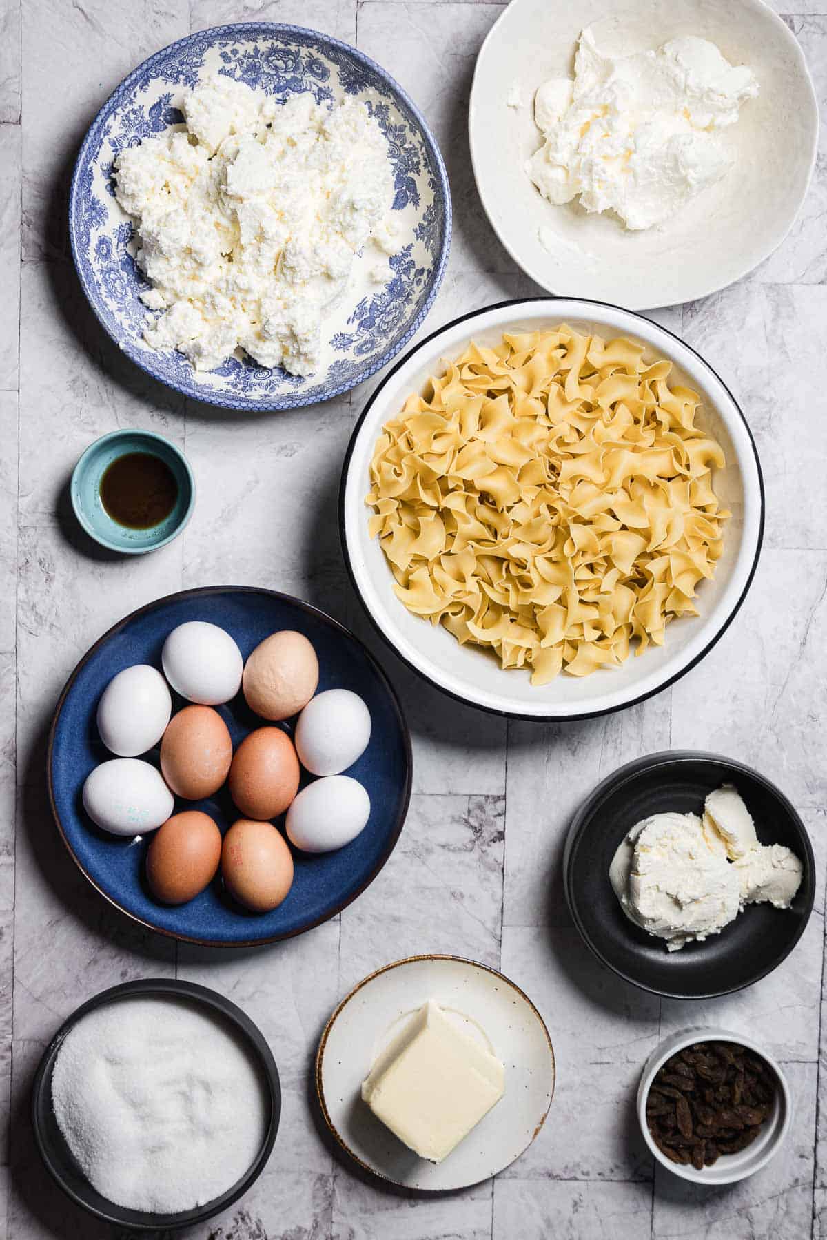 Ingredients for a sweet noodle kugel are neatly arranged on a marble countertop, featuring a bowl of uncooked egg noodles, a plate of eggs, and small bowls of sugar and raisins. Plates showcase ricotta cheese, sour cream, cream cheese, butter, and a dish of vanilla extract.