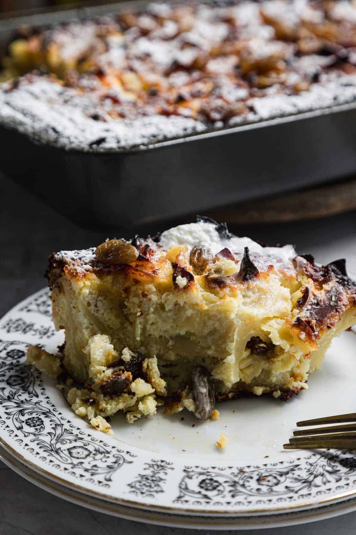 A slice of bread pudding with raisins is served on a patterned plate, alongside a fork. The golden brown pudding, dusted with powdered sugar, offers a tempting allure. In the background, a larger dish reminiscent of sweet noodle kugel completes this comforting scene.