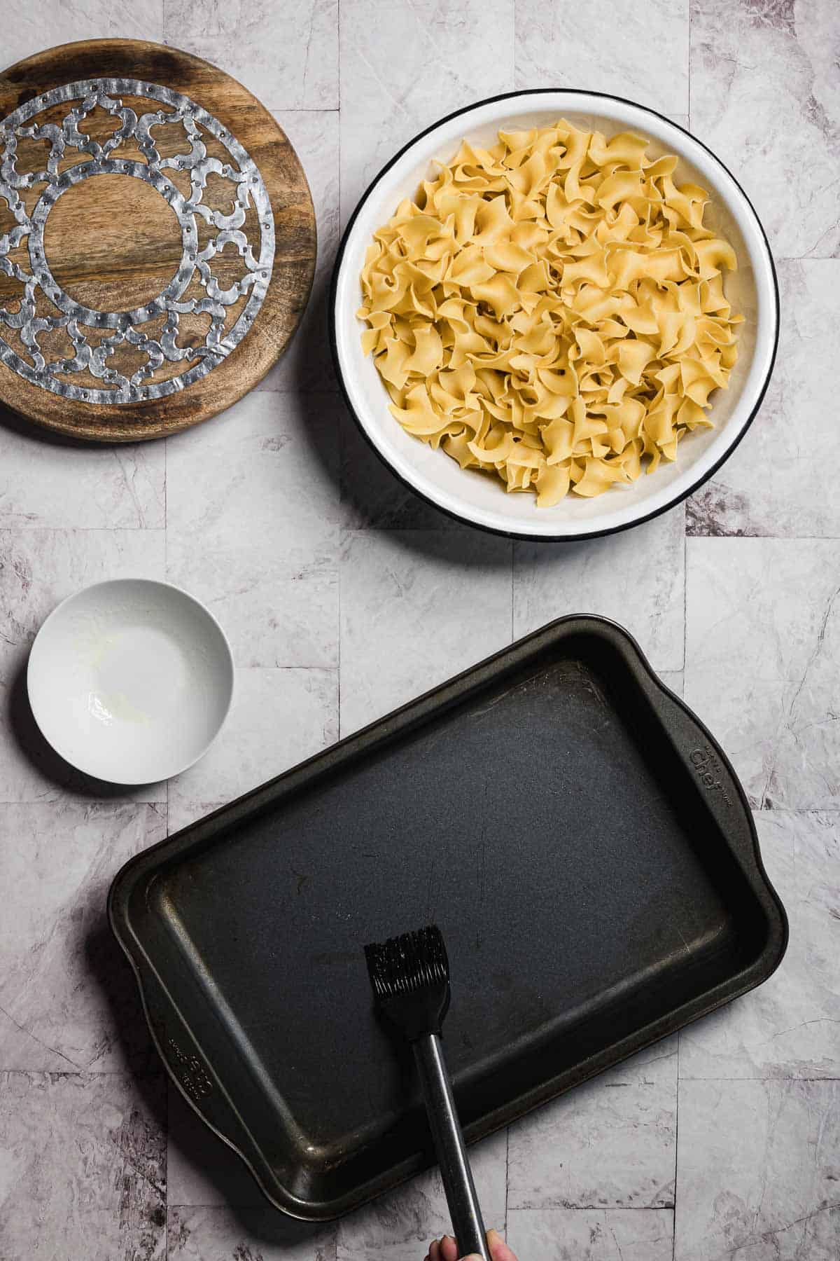 A marble surface showcases a round decorative wooden board, a bowl of uncooked pasta, hinting at a sweet noodle kugel, alongside a small white bowl and a dark rectangular baking tray. A hand holds a brush over the tray, poised to apply oil or butter.