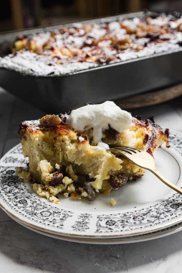 A slice of bread pudding, reminiscent of a sweet noodle kugel, is topped with whipped cream on a decorative plate. A fork is inserted into the dessert. In the background, a tray holds the rest of this delightful bread pudding treat.