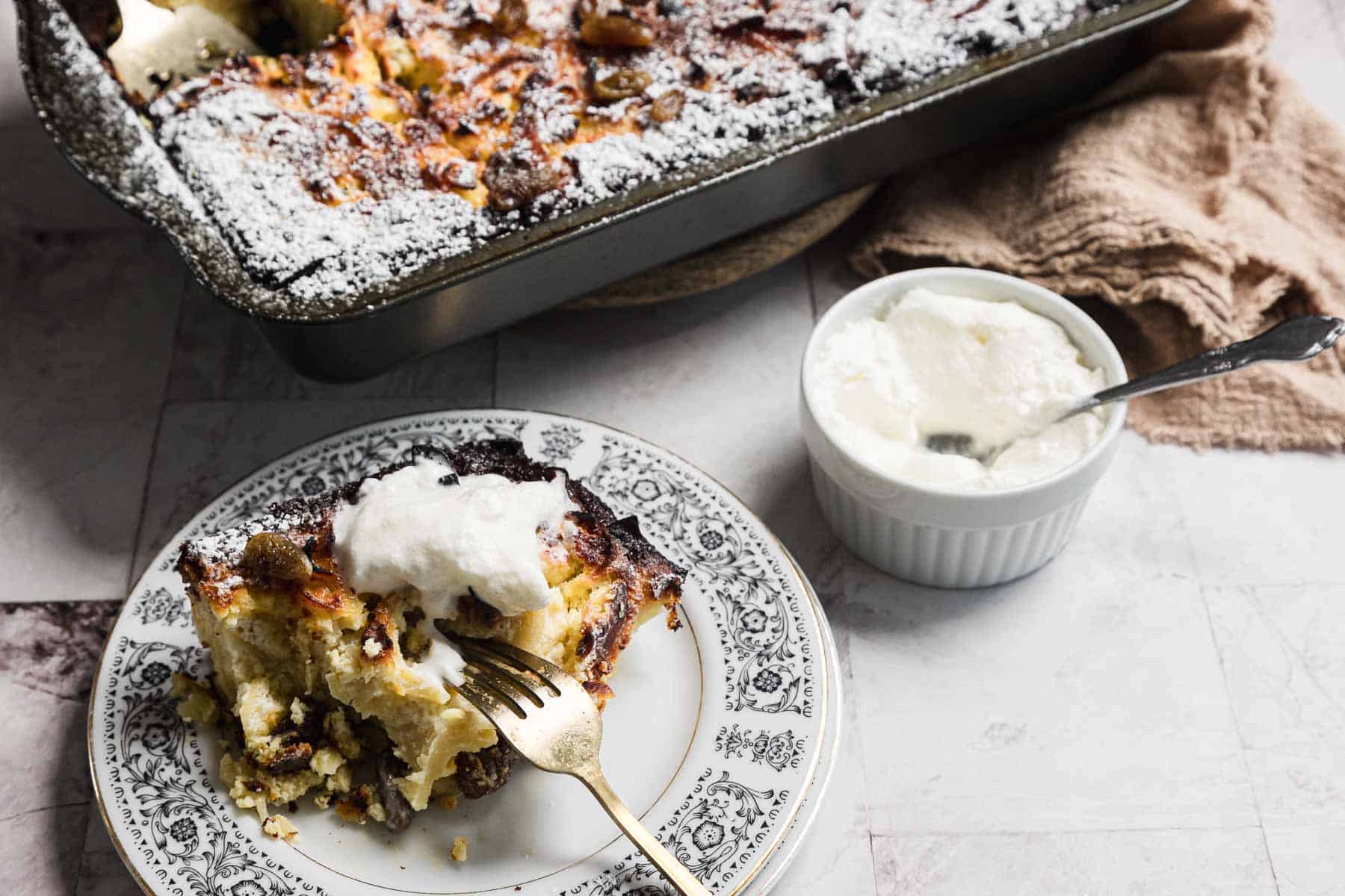 A piece of bread pudding on a decorative plate with a fork, topped with a dollop of whipped cream, sits invitingly. In the background, the baking dish hints at sweet noodle kugel inspiration. A small bowl with extra whipped cream and a spoon is beside the plate.