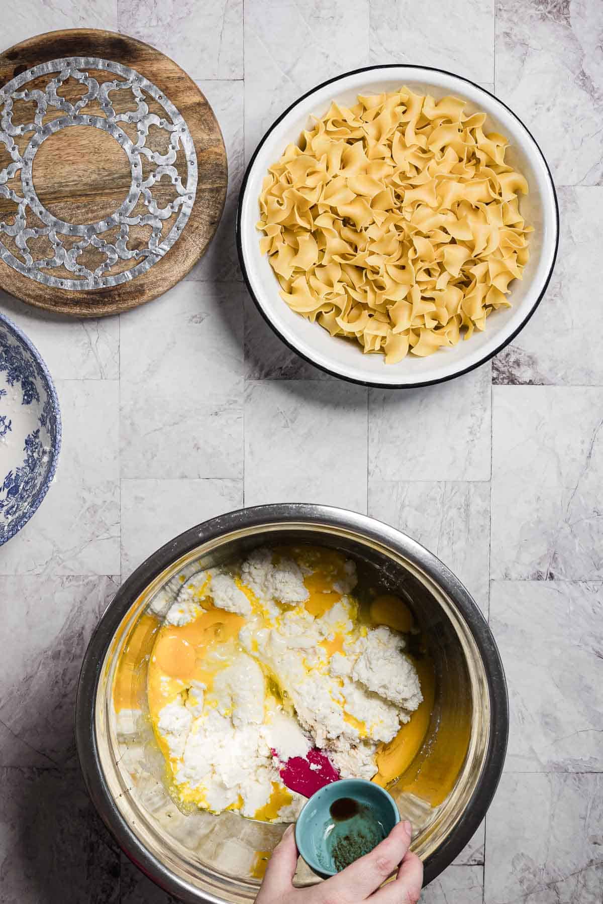 A kitchen counter displays a bowl filled with uncooked pasta noodles, ready for a sweet noodle kugel. A mixing bowl beneath it contains ingredients like eggs and ricotta cheese. A hand is adding liquid from a small cup while a cutting board and decorative plate sit nearby, waiting to assist in the creation.