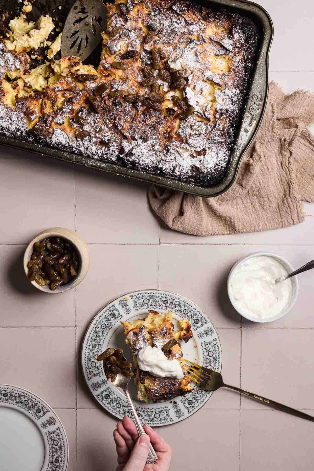 A baked sweet noodle kugel with a golden-brown crust sits in a black tray alongside a serving spatula. Below, a piece graces a decorative plate with fork and spoon, adorned with whipped cream. Nearby, a small bowl of sliced mushrooms and another of whipped cream await their turn.