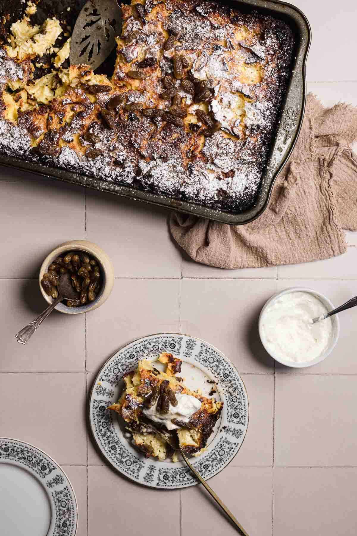 A baking dish filled with a browned sweet noodle kugel, sprinkled with powdered sugar. A plate beside it holds a serving topped with cream and nuts. A small bowl contains additional nuts, perfect for garnishing. A cloth is placed under the dish for an elegant touch.