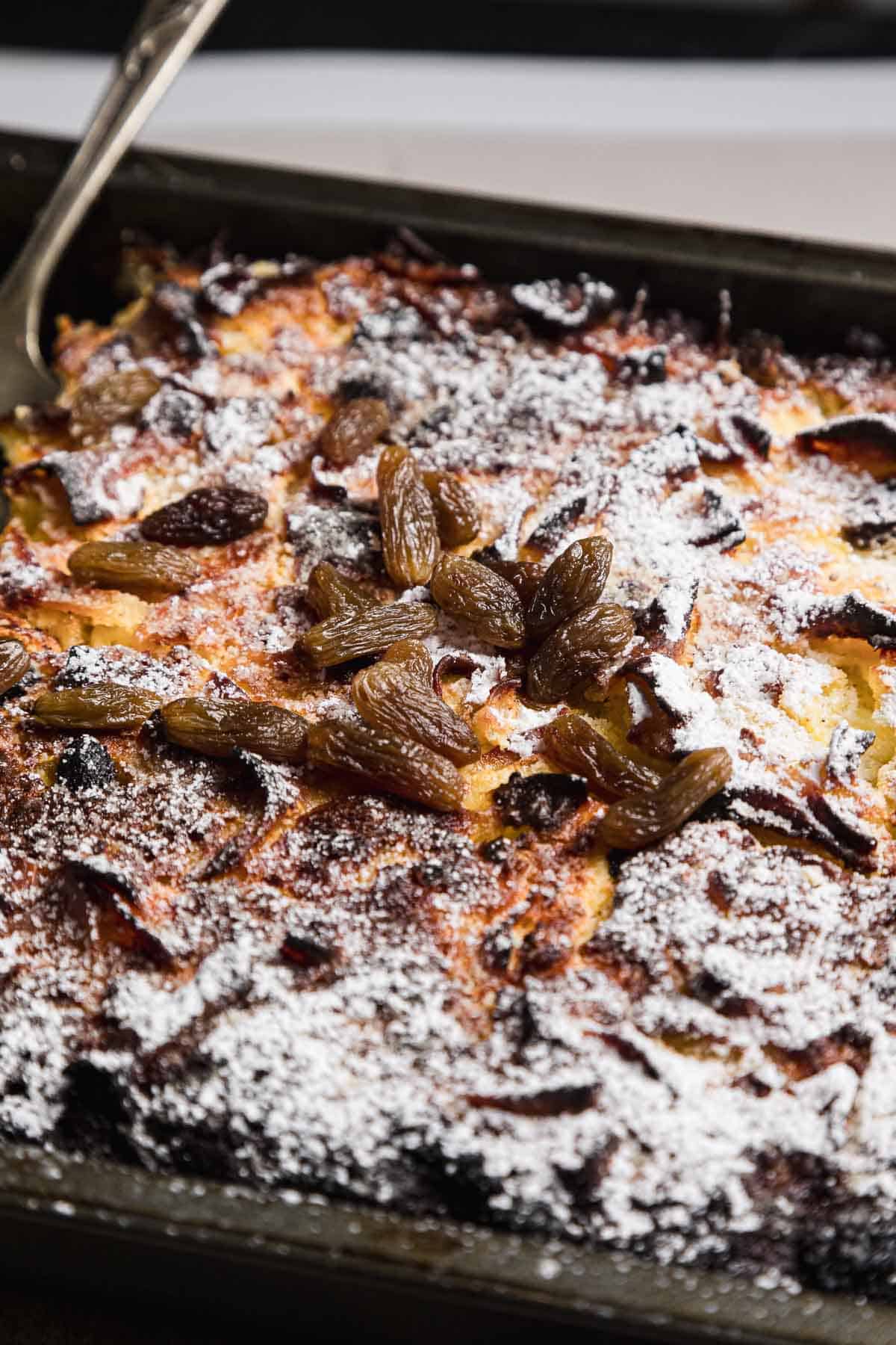 A close-up of a sweet noodle kugel topped with powdered sugar and raisins. The surface appears golden brown and slightly crispy. A portion of the dessert is nestled in a rectangular baking dish, with a serving utensil visible on the side, ready to serve this delightful treat.