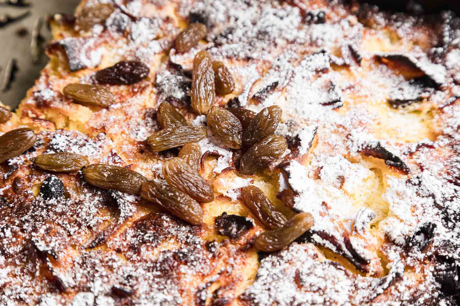 Close-up image of a baked sweet noodle kugel dessert topped with powdered sugar and golden raisins. The surface is browned and slightly crispy, featuring a delightful mixture of textures with scattered raisins throughout.