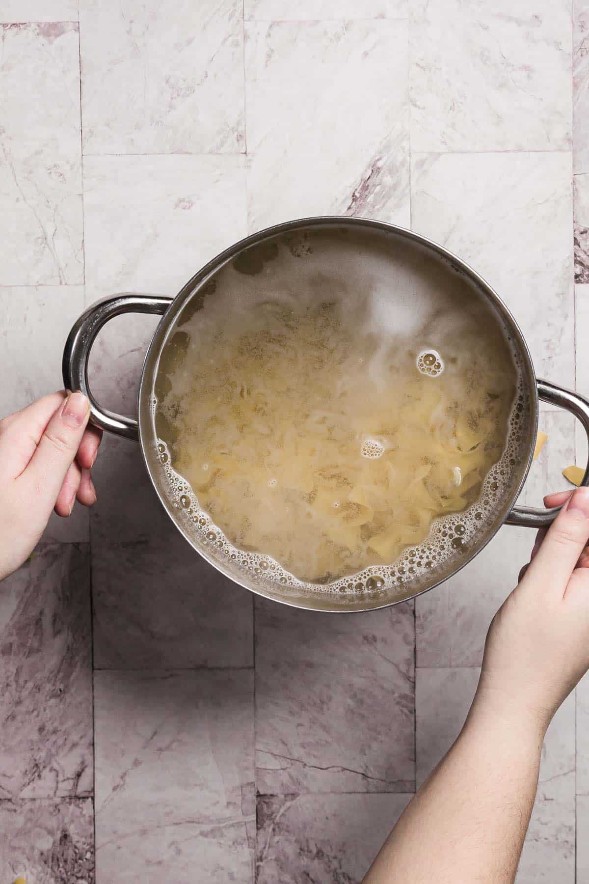 Hands hold a pot of boiling pasta, reminiscent of preparing sweet noodle kugel, with visible steam rising over a gray tiled countertop.